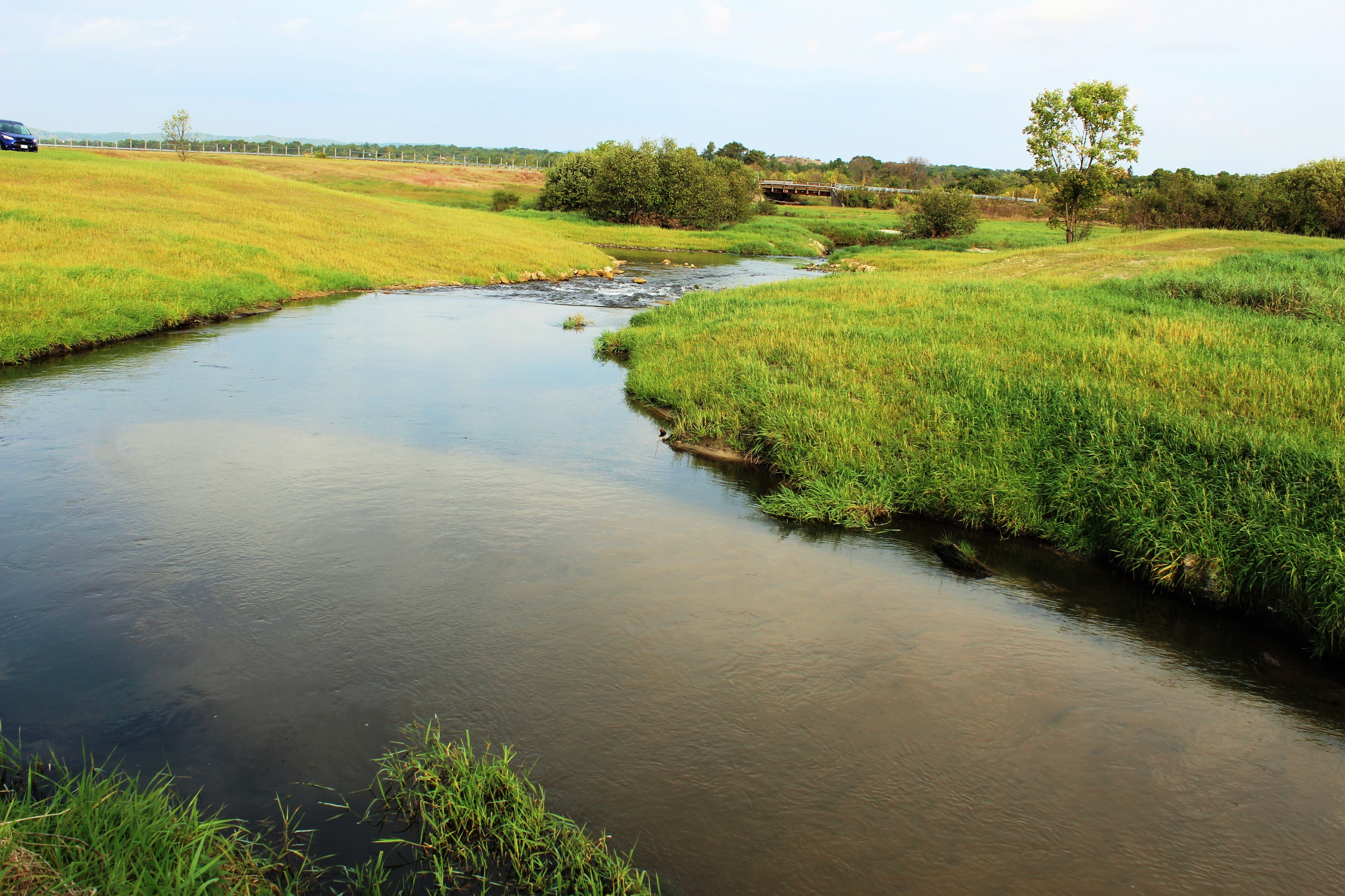 Photo of a creek in a wetlands area with green fields on both sides and a road and small bridge in the distance.