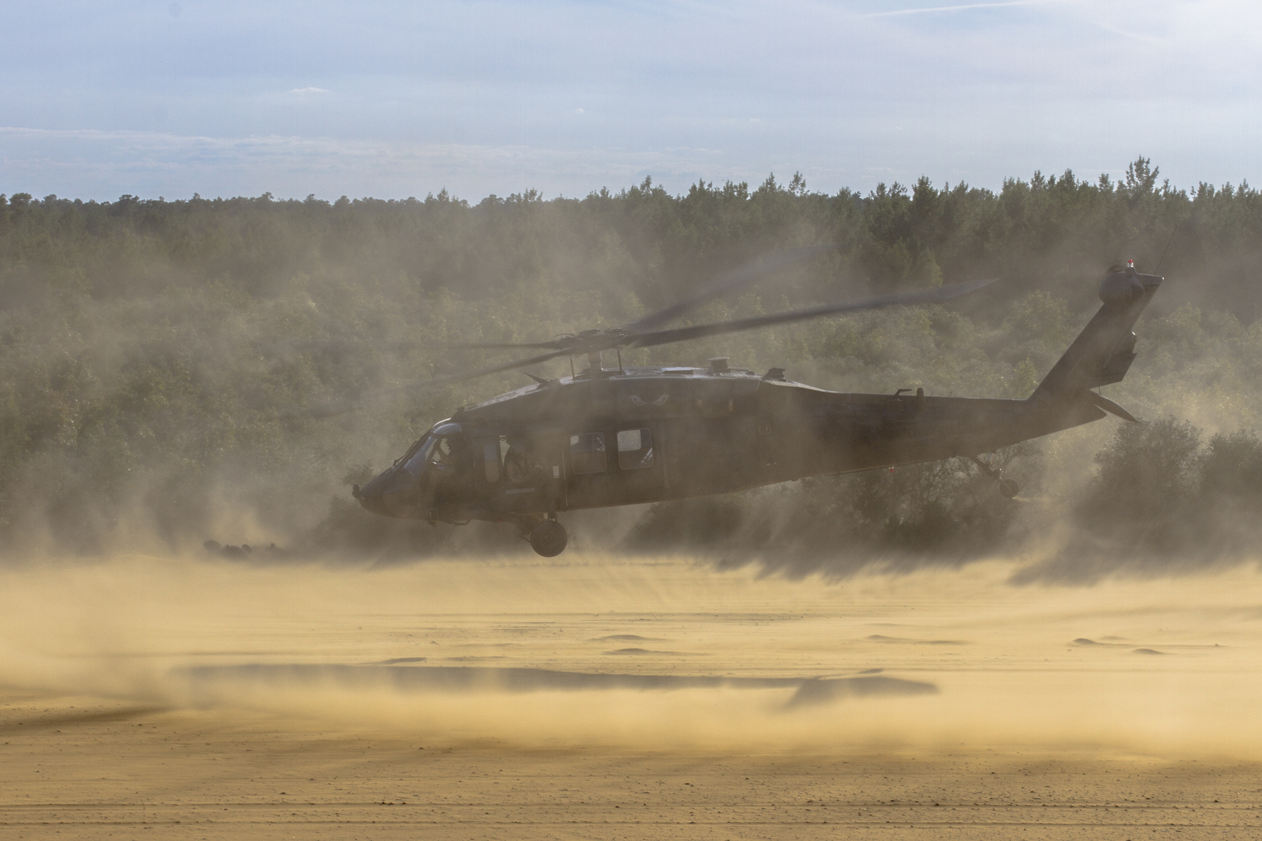 Photo of Army helicopter kicking up a lot of dust over a brown dirt area with a forest in the background.