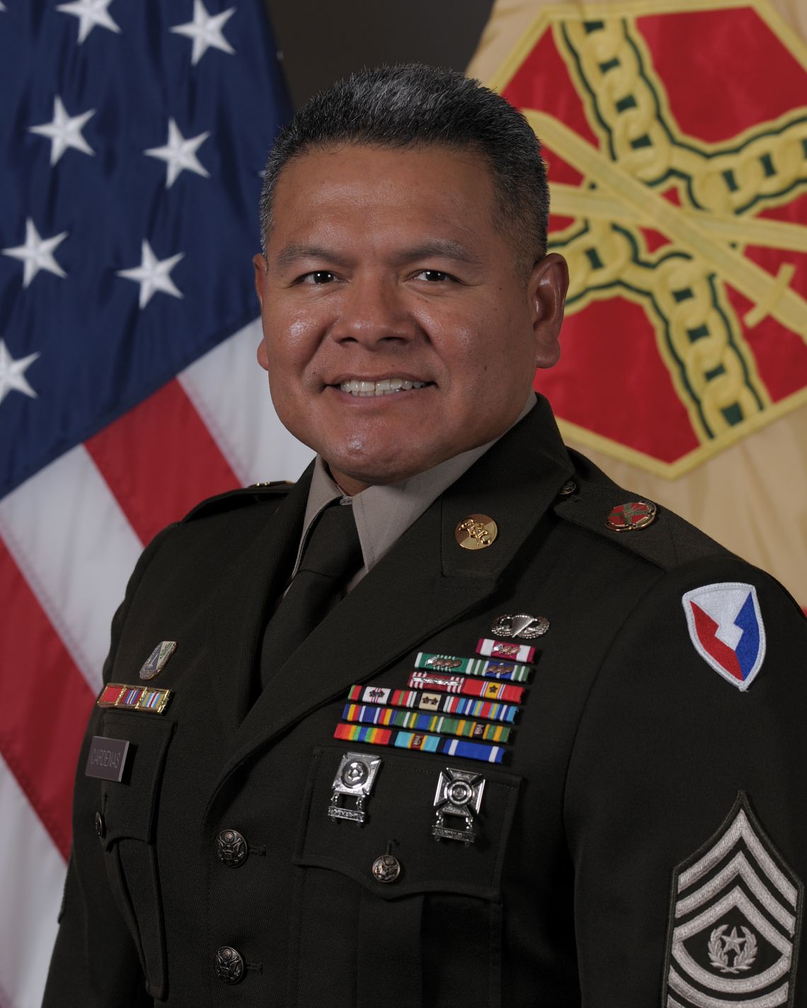 Photo of Command Sergeant Major Francisco Cardenas, Jr., in uniform standing in front of a U.S. and IMCOM flag.i